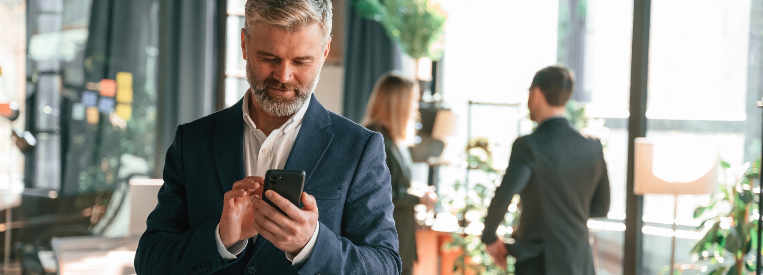 Ein Mann in einem dunkelblauen Anzug mit weißem Hemd steht in einem modernen Büro und hält ein Smartphone in beiden Händen. Im Hintergrund sind zwei weitere Personen zu sehen, die sich unterhalten. Die Umgebung ist hell und mit großen Fenstern ausgestattet.