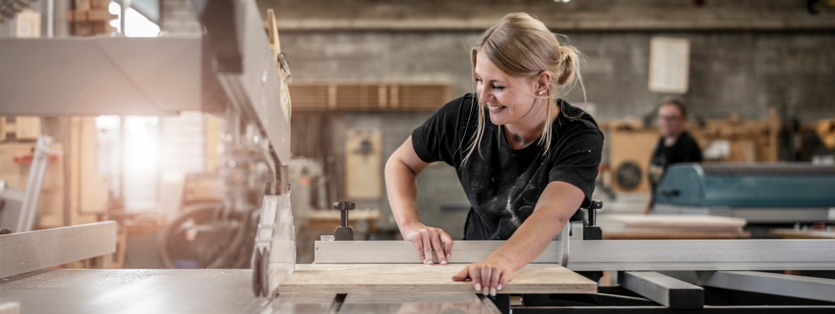 Das Bild zeigt eine blonde Frau, die in einer Werkstatt mit einer Tischkreissäge arbeitet und ein Holzbrett zuschneidet.  Im Detail zeigt das Bild eine junge Frau mit blonden, hochgesteckten Haaren, die ein dunkles T-Shirt trägt, das mit Holzstaub bedeckt ist. Sie steht an einer Tischkreissäge und führt ein helles Holzbrett mit konzentriertem Blick und einem leichten Lächeln dem Sägeblatt entgegen. Ihre Hände sind sorgfältig positioniert, um das Holz sicher zu führen. Die Säge selbst ist groß und robust, mit einer grauen Schutzvorrichtung über dem Sägeblatt. Im Hintergrund ist eine andere Person verschwommen zu erkennen, ebenfalls in dunkler Kleidung, was darauf hindeutet, dass es sich um eine Werkstattumgebung handelt. Der Raum ist gut beleuchtet, mit natürlichem Licht, das durch Fenster einfällt und eine warme und einladende Atmosphäre schafft. Die Stimmung des Bildes ist positiv und vermittelt ein Gefühl von Handwerkskunst, Präzision und Freude an der Arbeit.